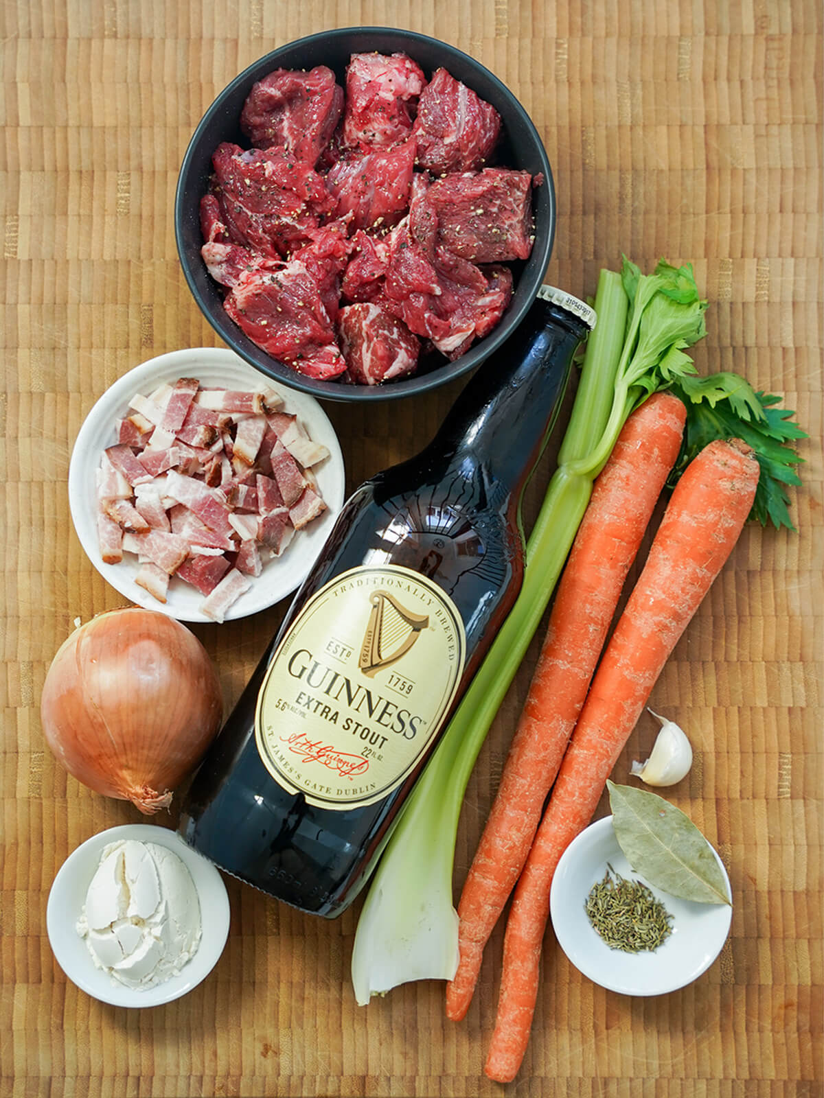 bottle of Guinness, bowl of beef chunks, carrots, onion, celery, dish with chopped bacon and small dishes with herbs and flour.