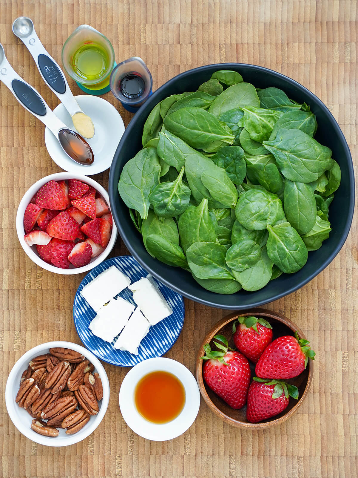 bowl of spinach, bowls with strawberries, chunks of feta, pecans and measures with syrup, honey, oil and vinegar.