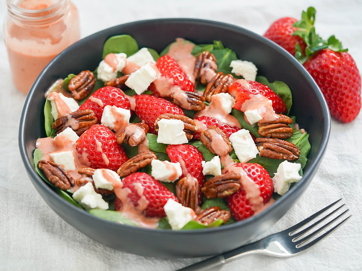 side view of bowl of spinach and strawberry salad with fork in front and jar of remaining dressing and strawberries behind bowl.
