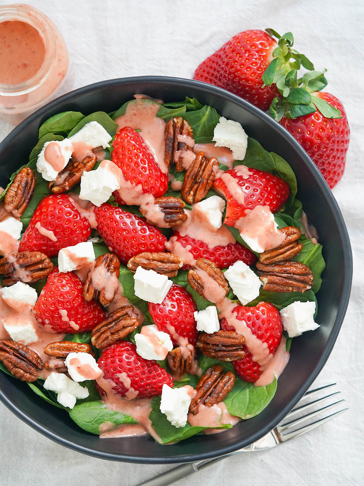 bowl of spinach and strawberry salad with fork below bowl, strawberries above, viewed from overhead.