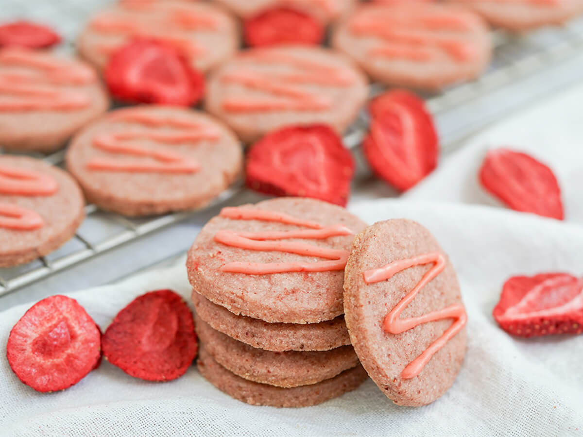 strawberry shortbread cookies small stack of strawberry shortbread cookies with freeze dried strawberry pieces around and more cookies behind.