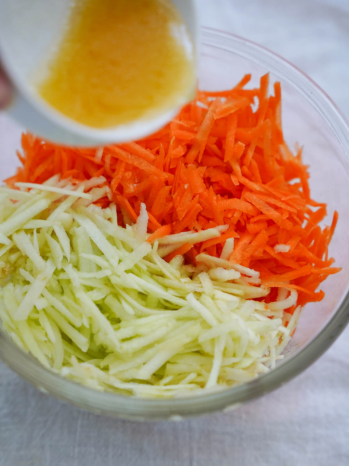 grated carrot and apple in a bowl, with small dish above, pouring dressing over.