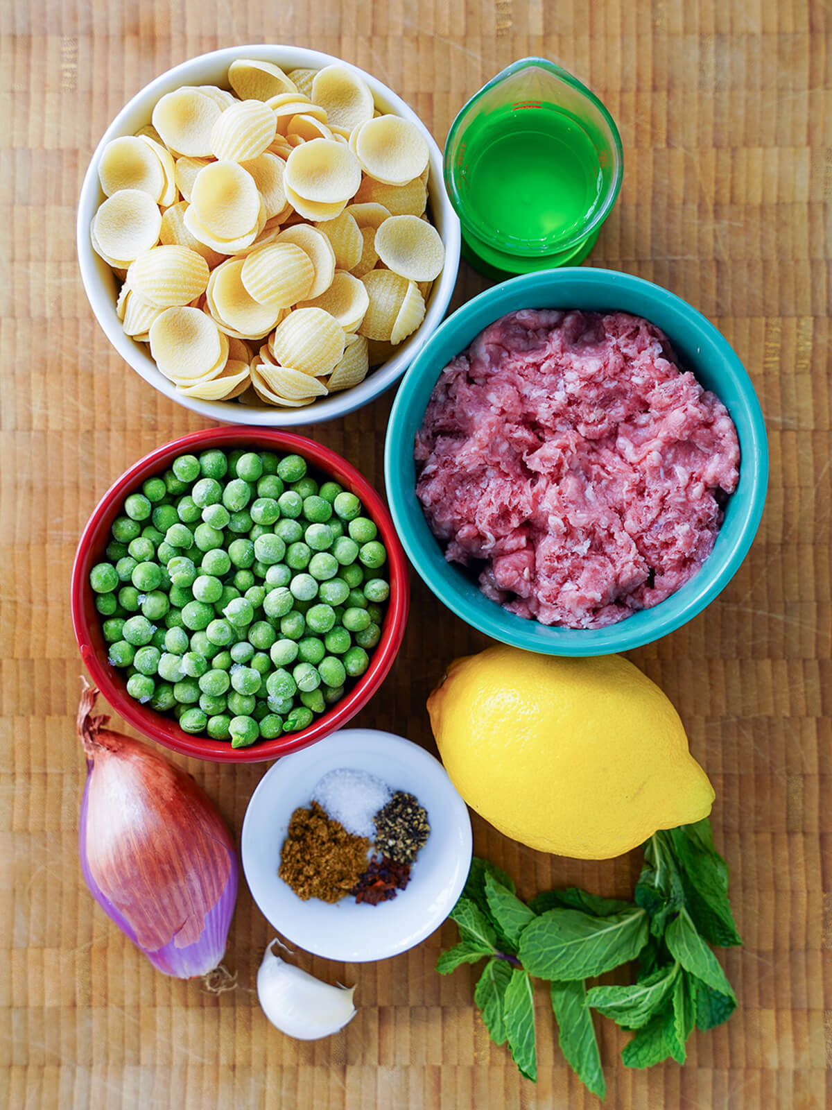 bowls of dried pasta, ground lamb, peas, with a lemon, dish with spices, mint sprig, shallot, garlic.