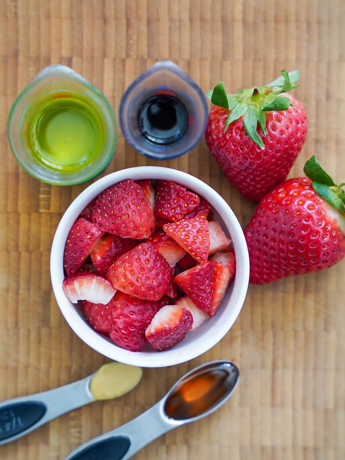 overhead view of a bowl of strawberries with measures of oil, balsamic, spoons of honey and mustard around bowl.
