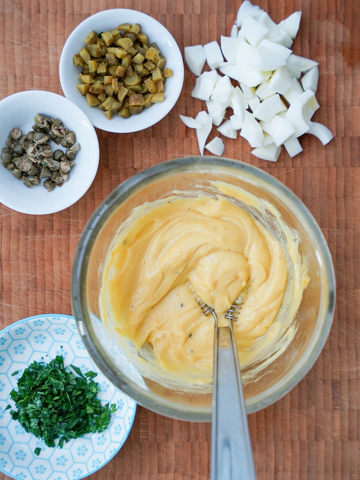 bowl of egg-oil emulsion with chopped egg white, chopped cornichons, capers and chopped parsley in dishes to side.