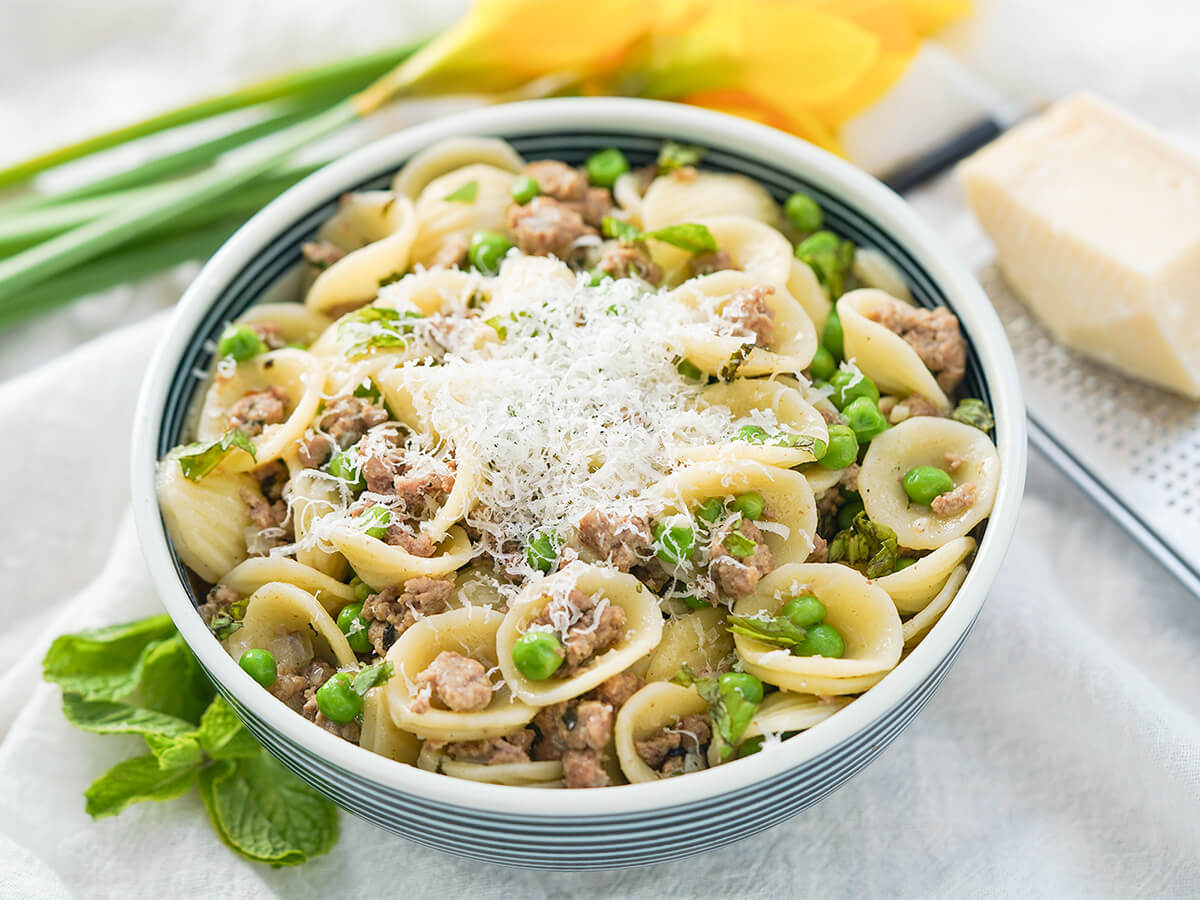bowl of pea and lamb pasta topped with cheese with block of cheese on grater behind.