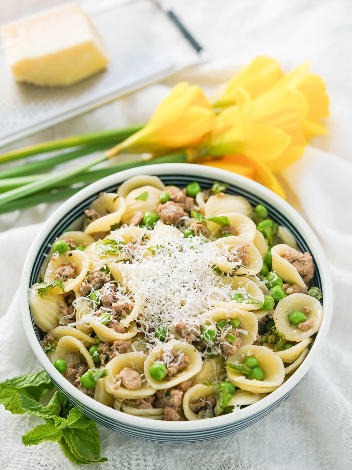 bowl of pea and lamb pasta with bunch of daffodils and grater with cheese in background.