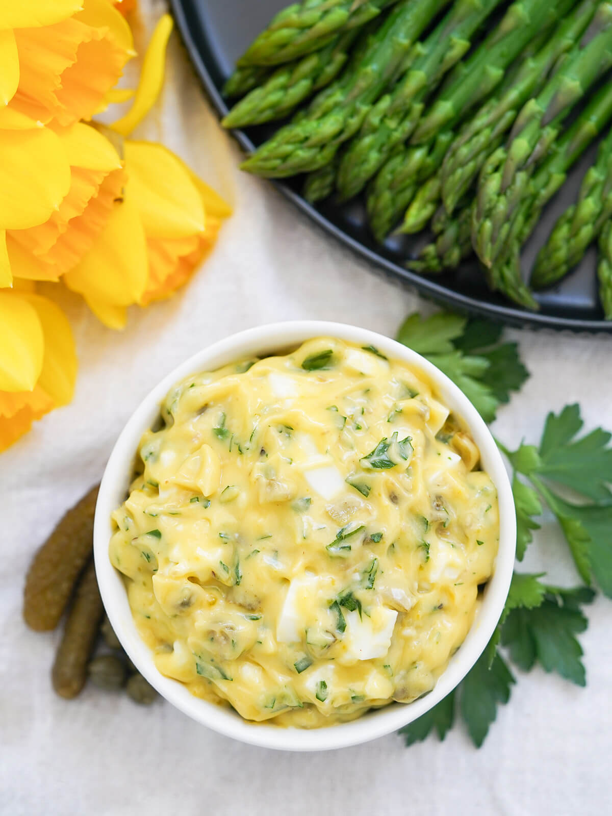 overhead view of a dish of sauce gribiche with part view of plate of asparagus and bunch of daffodils above.
