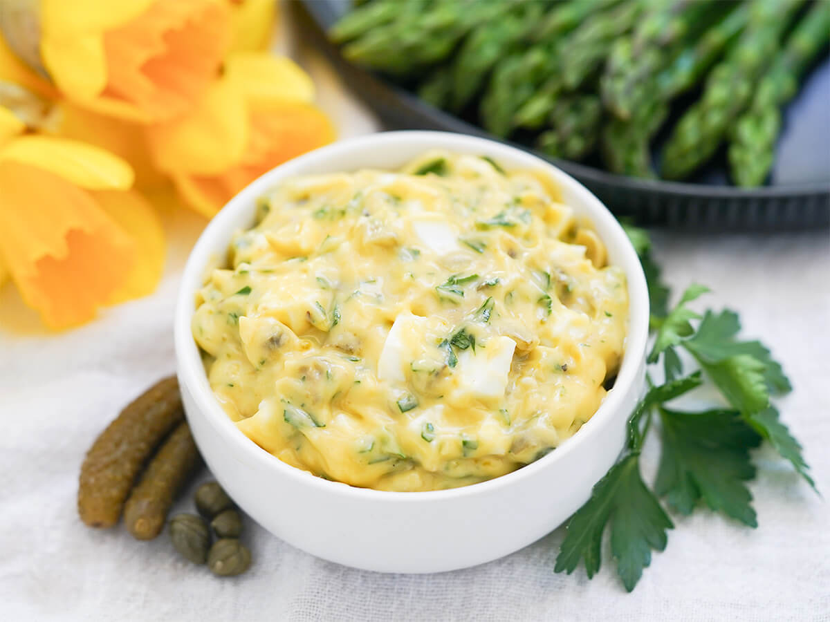dish of sauce gribiche with parsley, cornichons and capers to sides of bowl and asparagus and daffodils blurred behind.