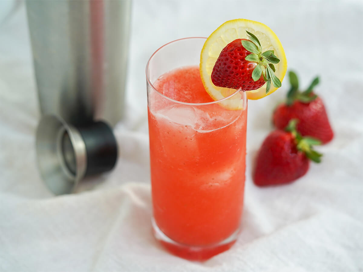 glass of strawberry collins garnished with slice of lemon and a strawberry with strawberries behind glass.