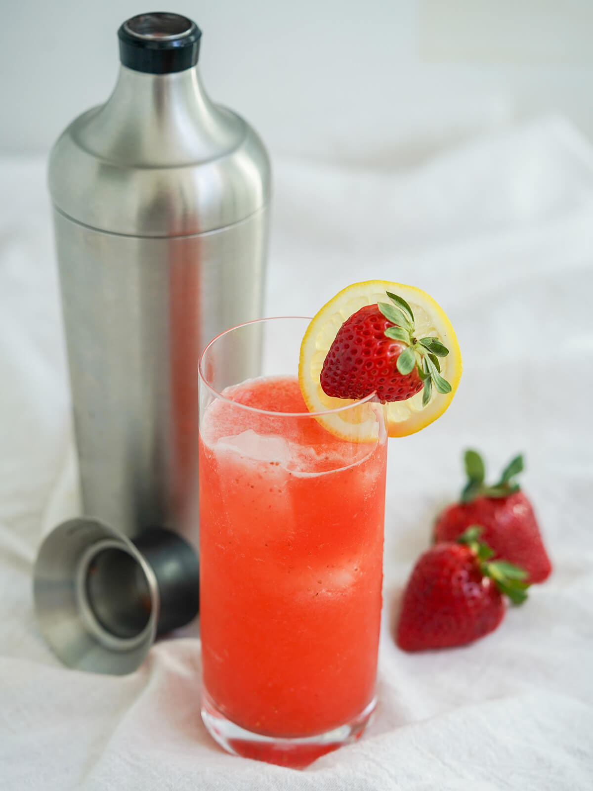 glass of strawberry collins next to cocktail shaker with strawberries to side.