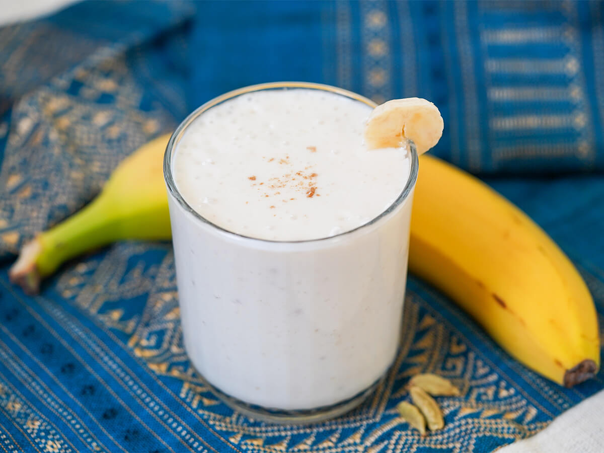 glass of banana lassi with banana behind glass and cardamon pods to side.