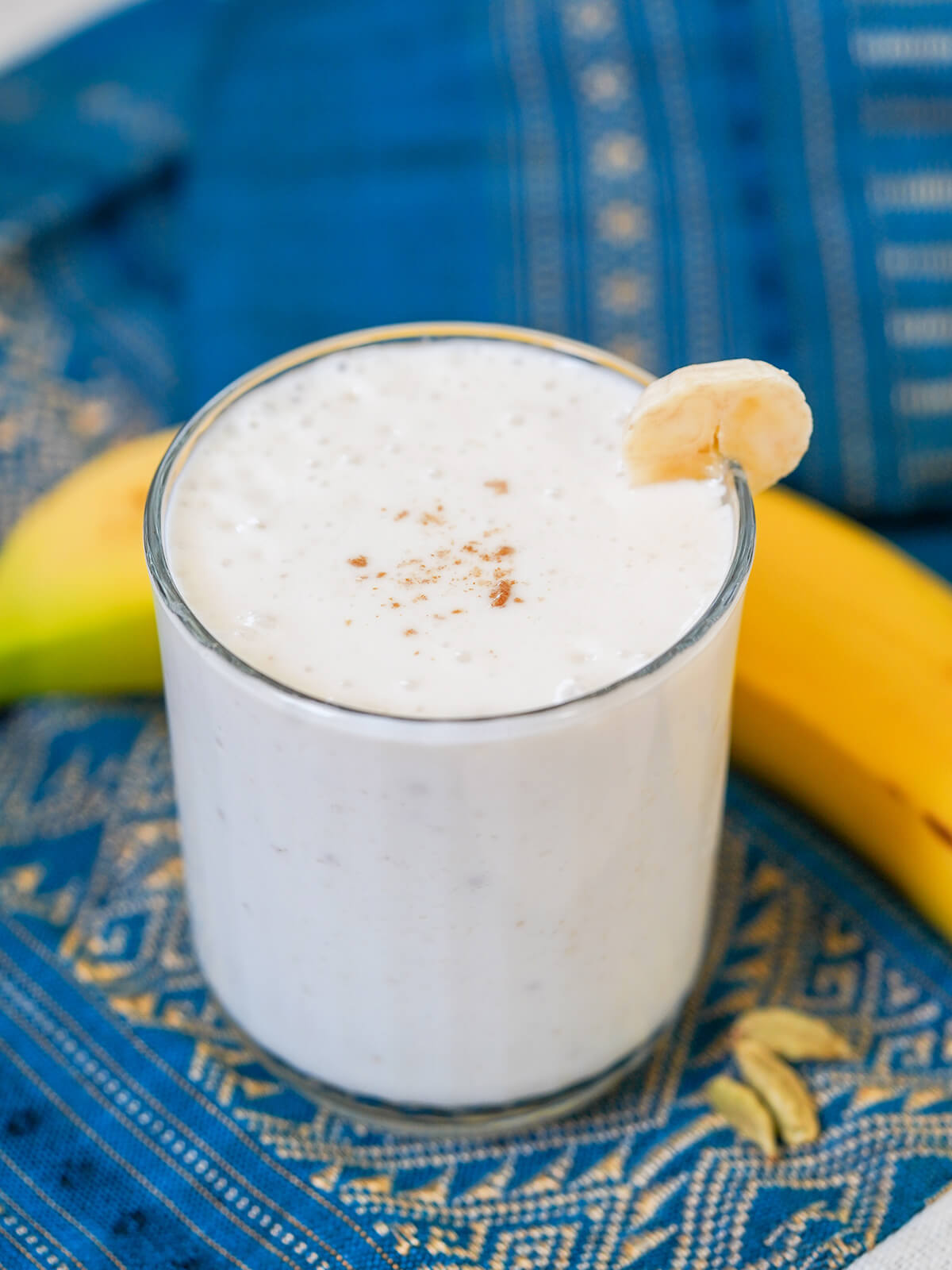 side view of glass of banana lassi with slice of banana on glass.