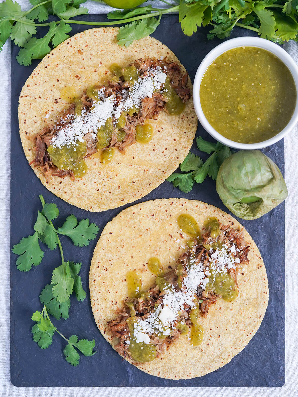 overhead view of two tacos topped with pork carnitas, salsa verde and a little cheese with dish of salsa verde to side and cilantro around.