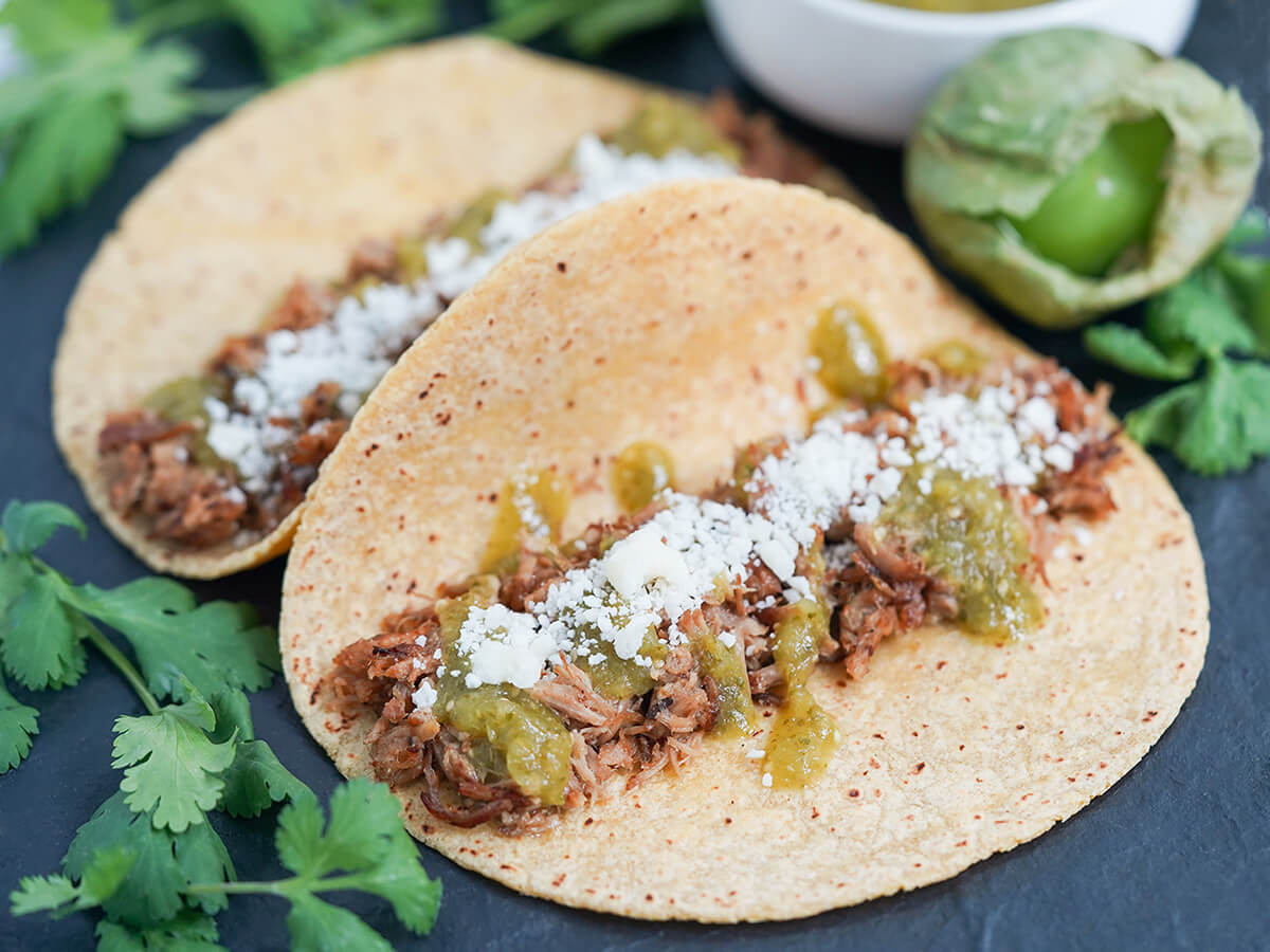 two carnitas tacos with salsa verde with cilantro either side and a tomatillo to side.