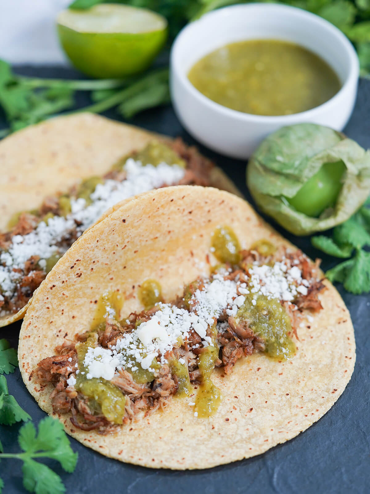 a taco with pork carnitas and salsa verde with more sauce and tomatillo behind and a second taco resting behind one at front.