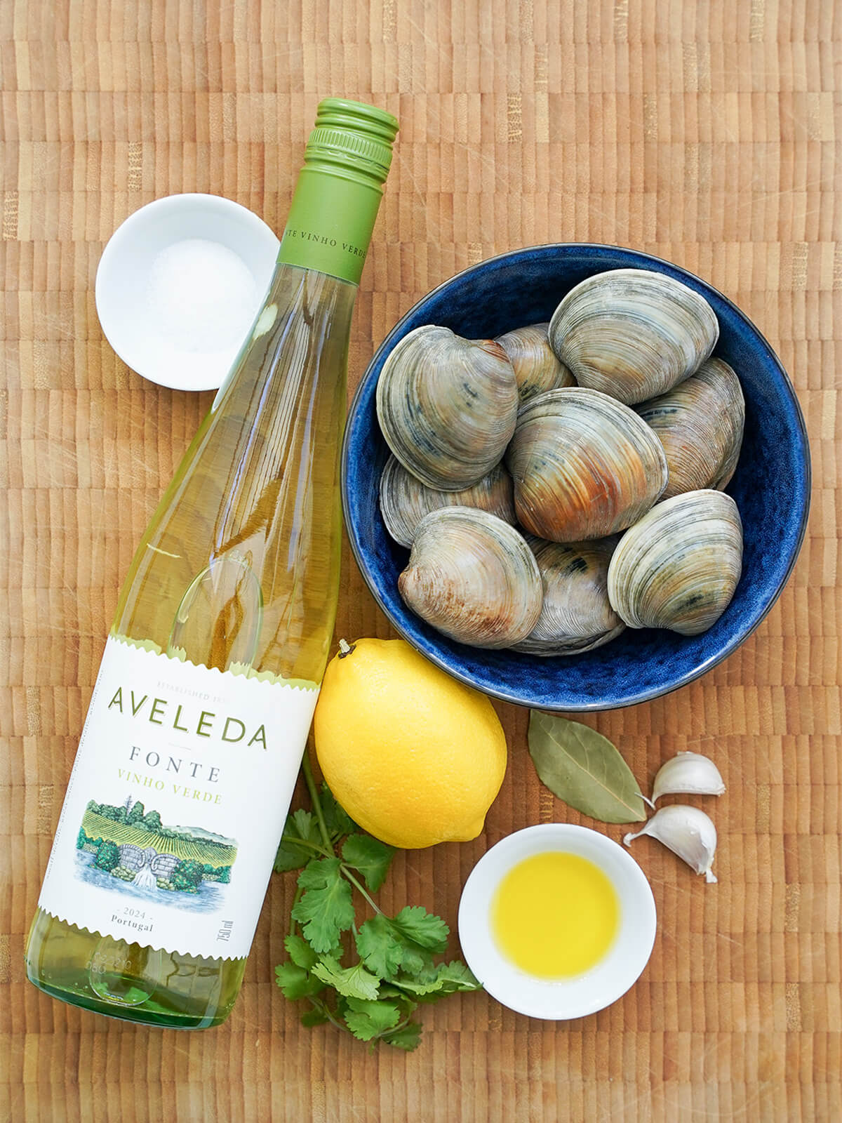 bowl of clams, lemon, bay leaf, garlic cloves, bottle of wine, dish with olive oil and sprig of cilantro.