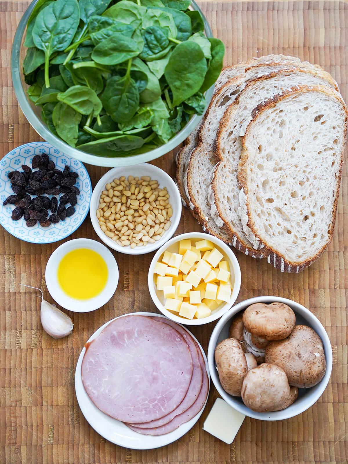 ingredients for llescas, Catalan loaded toasts bowl of spinach, small dishes with raisins, pine nuts, cubes of cheese, mushrooms and olive oil with stack of sliced bread to one side and garlic to other.