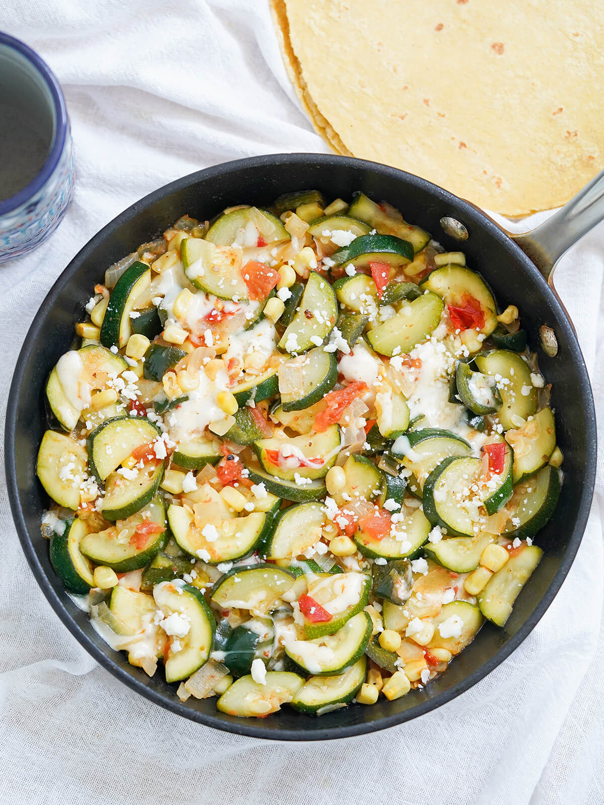 Calabacitas is an easy and delicious Mexican zucchini dish, with corn, tomato and cheese. It's quick, fresh yet comforting and perfect as a side dish, taco filling or vegetarian main. overhead view of skillet of calabacitas, Mexican zucchini with tortillas above.