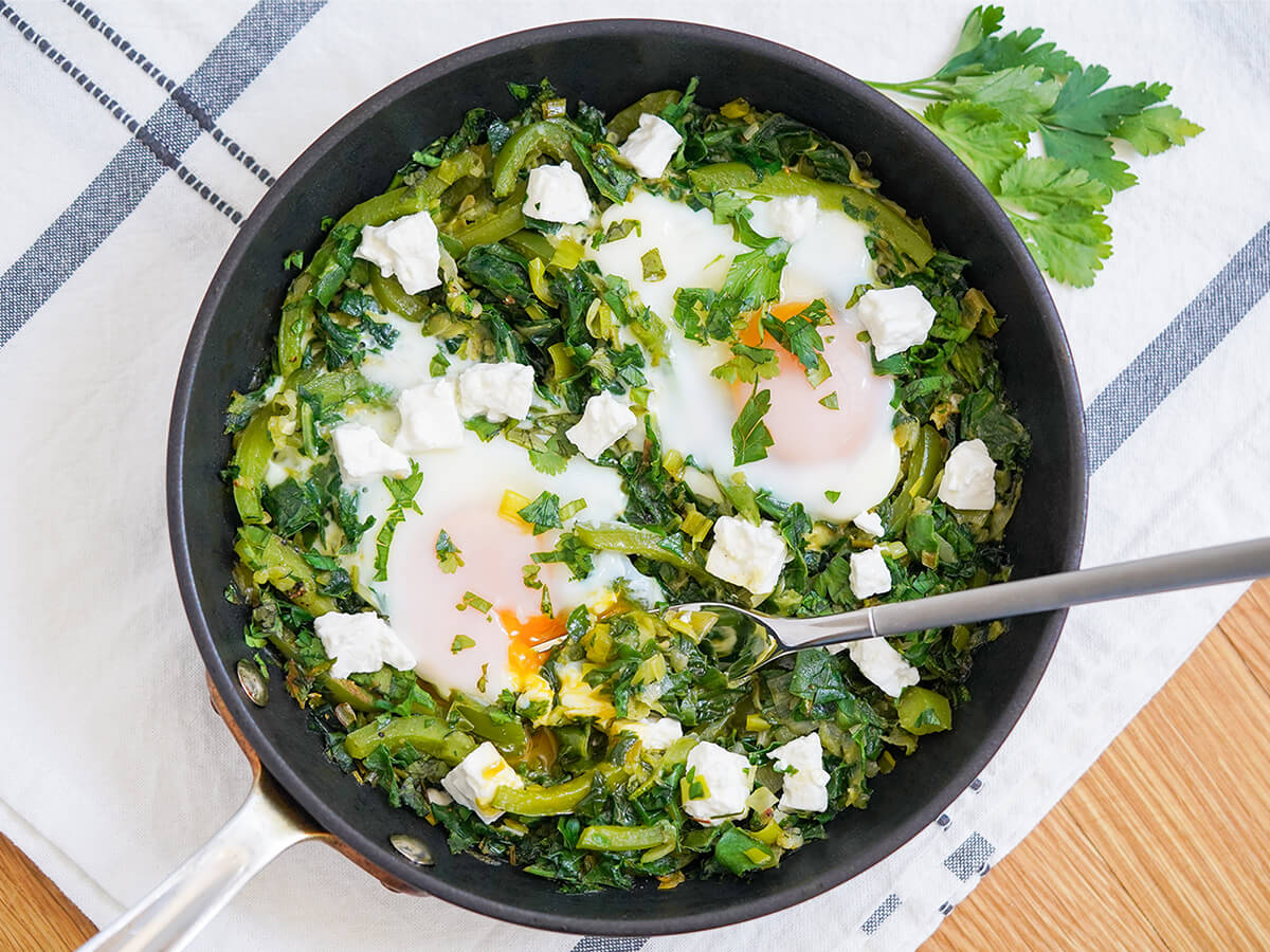 overhead view of skillet of green shakshuka with fork in skillet and herbs above skillet on cloth.