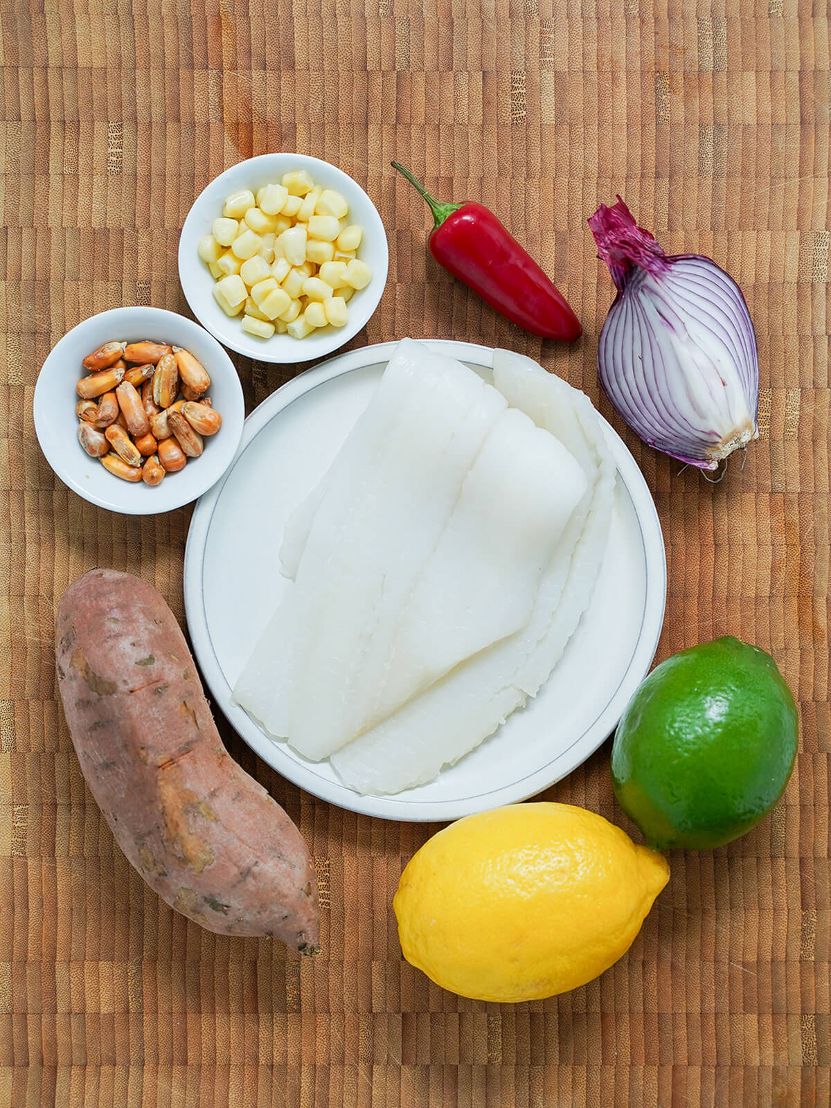 plate with pieces of white fish, with a sweet potato, lemon and lime, red onion wedge, chili and corn around plate.