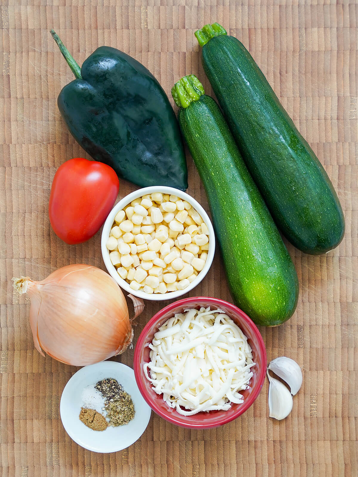 ingredients for calabacitas, Mexican zucchini zucchini, poblano pepper, tomato, onion, garlic cloves with bowls if corn and grated cheese in middle.