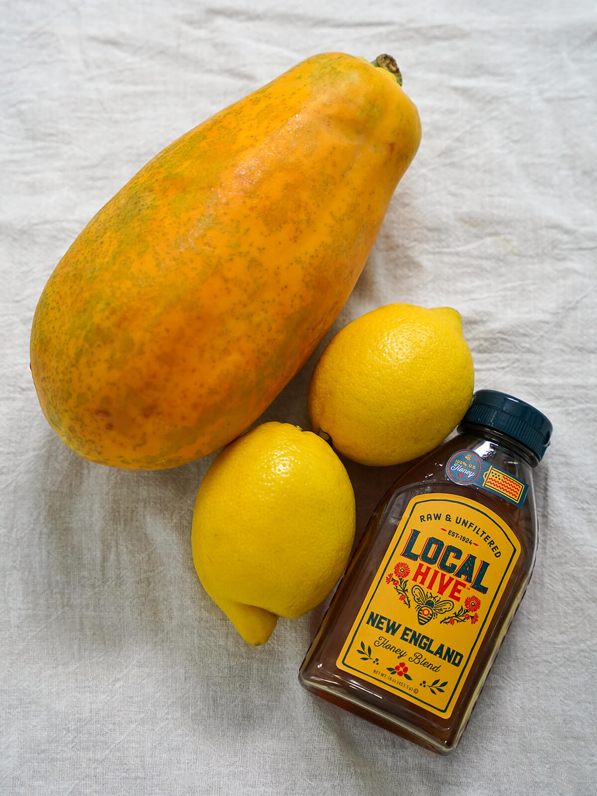 whole papaya, two lemons and a container of honey on cloth.