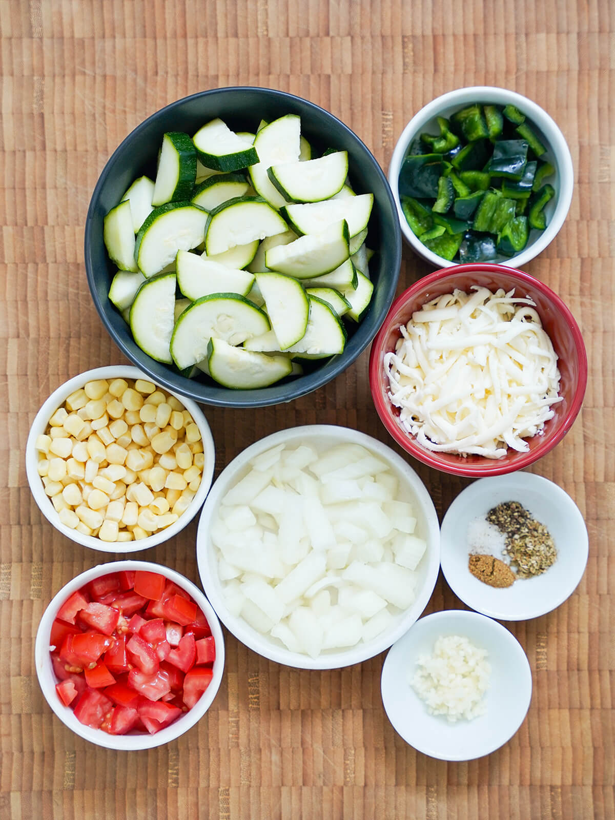 chopped up ingredients for calabacitas dishes of sliced zucchini, chopped pepper, onion, corn, tomato and shredded cheese with smaller dishes of spices and garlic.
