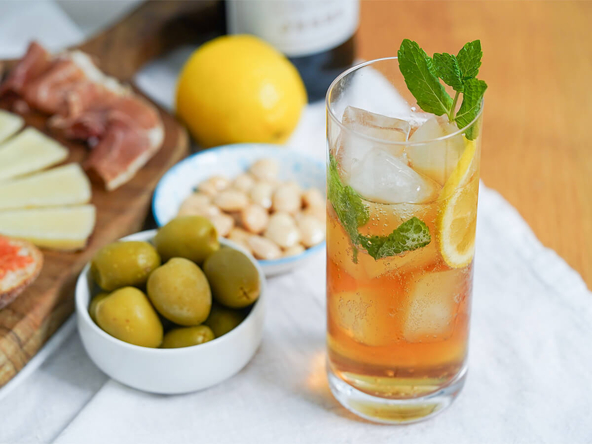 glass of rebujito, sherry cocktail with dishes of olives, almonds to side and lemon behind.