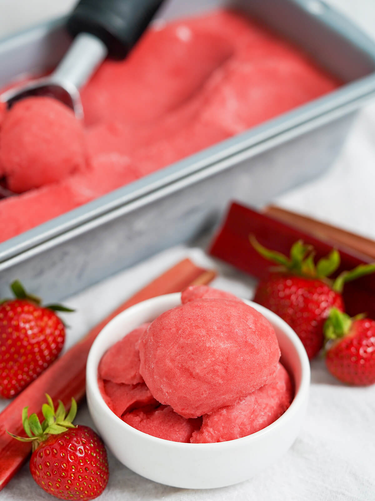 bowl of strawberry rhubarb sorbet with tin with more behind and scoop sitting on top.