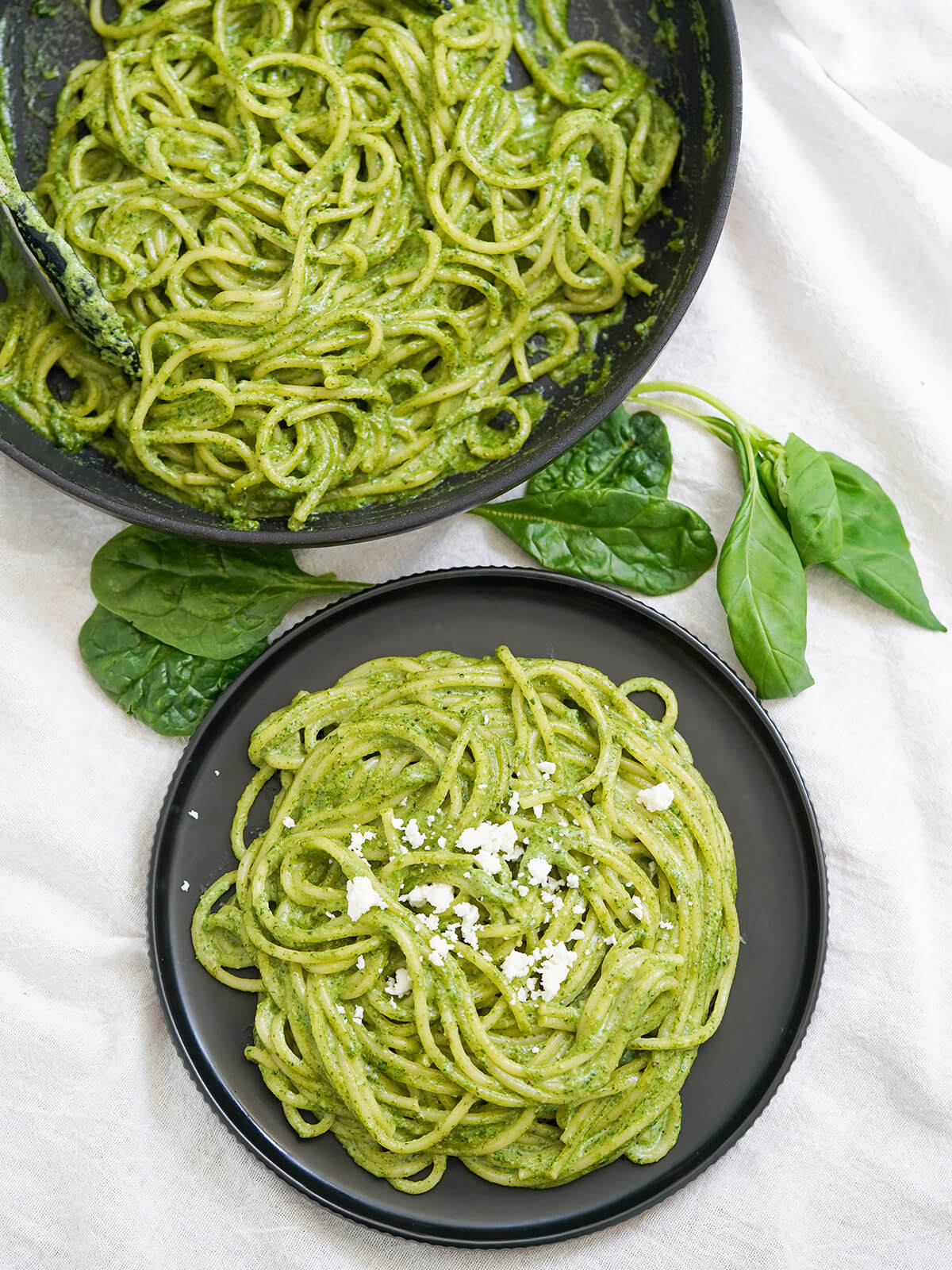 overhead view of plate of tallarines verdes, also called Peruvian pesto, with skillet with more in it above and spinach and basil leaves in between.