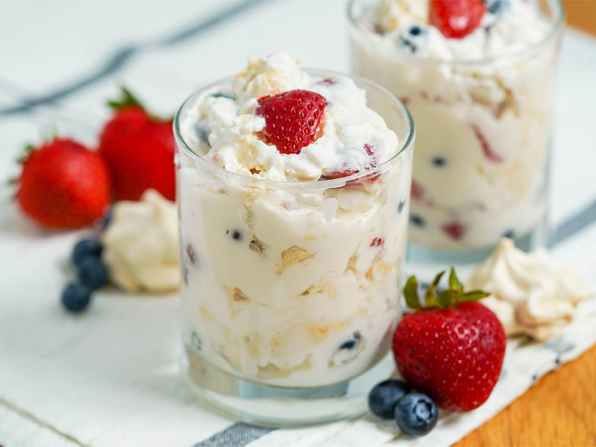 Eton mess is a classic British dessert that is incredibly easy to prepare - simply fruit, cream and meringue mixed together - but the result is so delicious. It's perfect for a party or easy anytime treat. side view of a glass of Eton mess with strawberries and blueberries to side and behind.