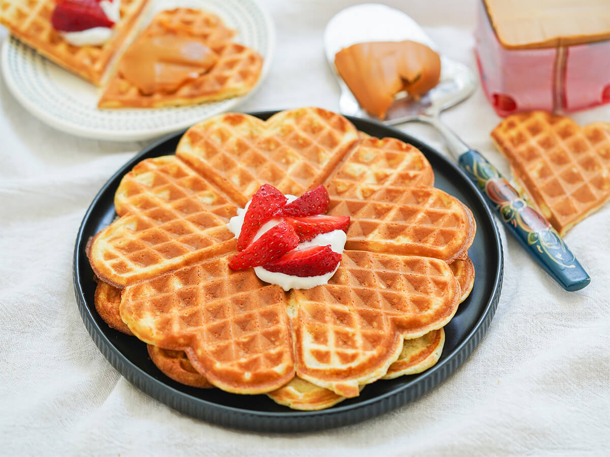 two Norwegian waffles stacked on a plate with heart-shaped pieces behind and to side, cheese spar with slice of brown cheese and block of cheese to side.