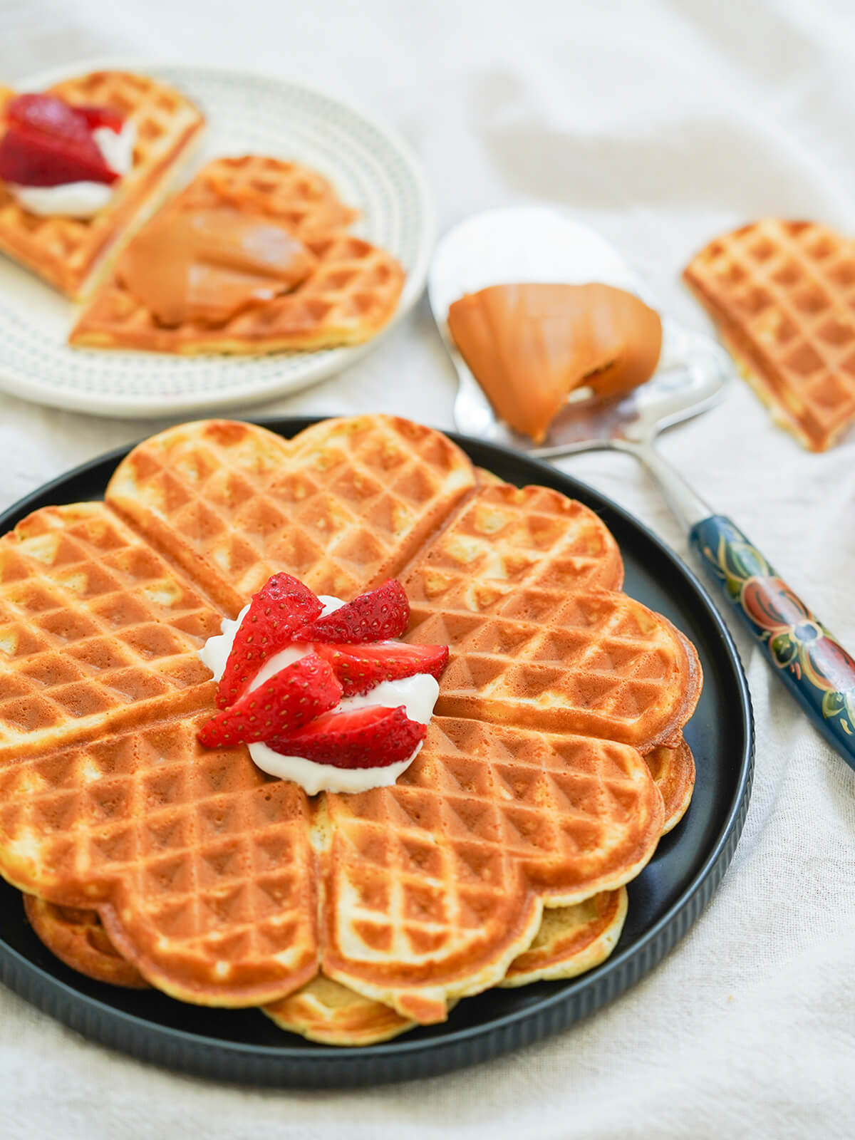 side view of Norwegian waffles, vafler, on plate with cheese spar topped with brown cheese behind and some waffle wedges on small plate blurred in background.
