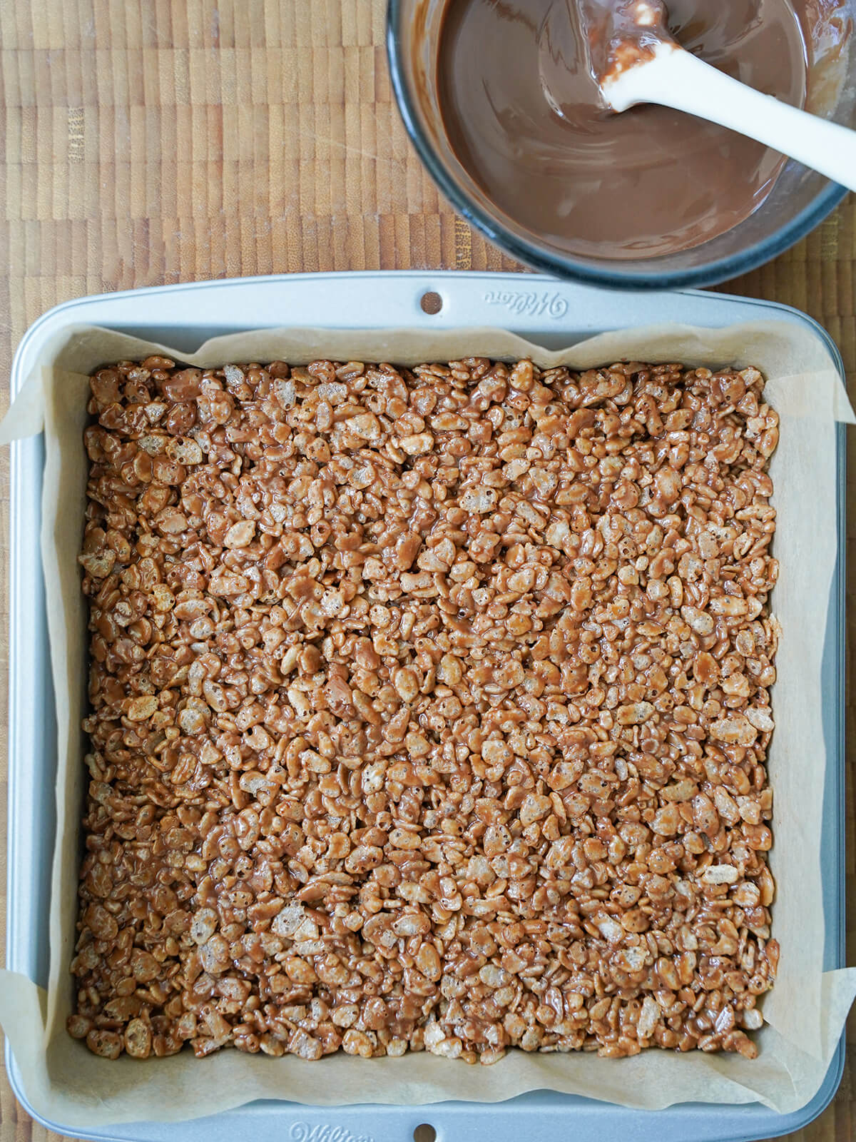 layer of candy-coated rice cereal in lined baking pan with bowl of melted chocolate above. coated rice krispie mixture in lined baking pan with bowl of melted chocolate above.