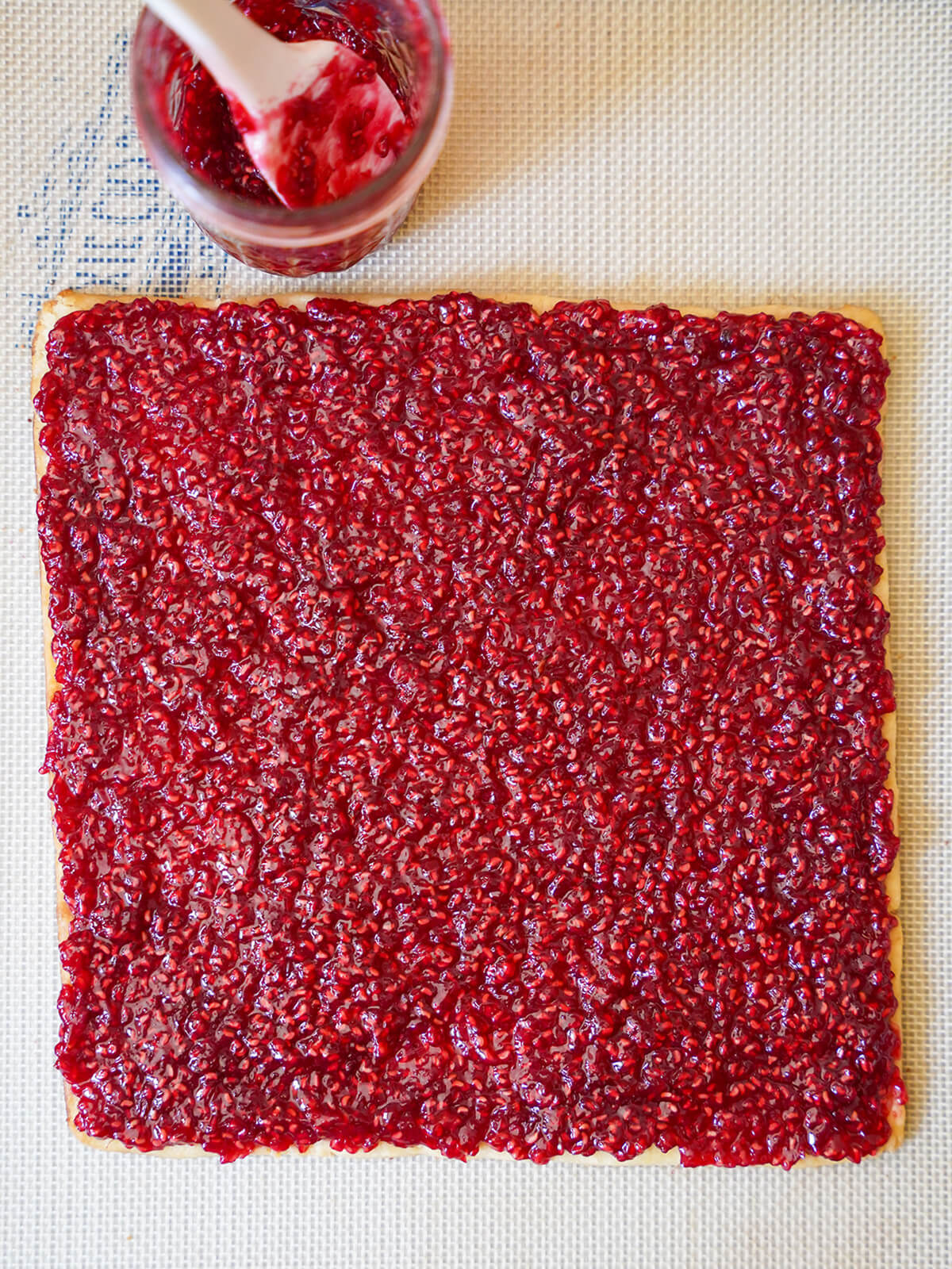 raspberry jam spread over a square of cookie with spatula in jar above.