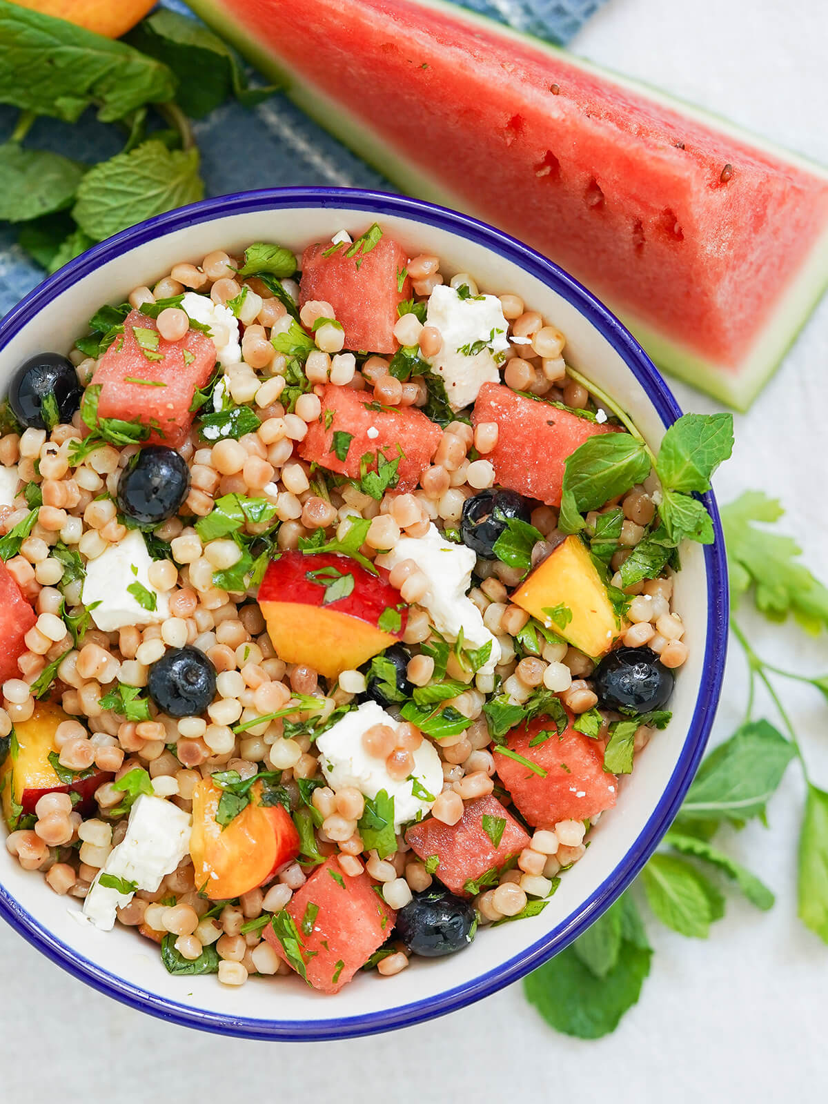 This fruit couscous salad with herbs and feta is light, bright and refreshing with juicy fresh fruit & aromatic herbs, but filling enough to be a great lunch or side. overhead part view of bowl of fruit couscous salad with herbs around bowl and wedge of watermelon above.