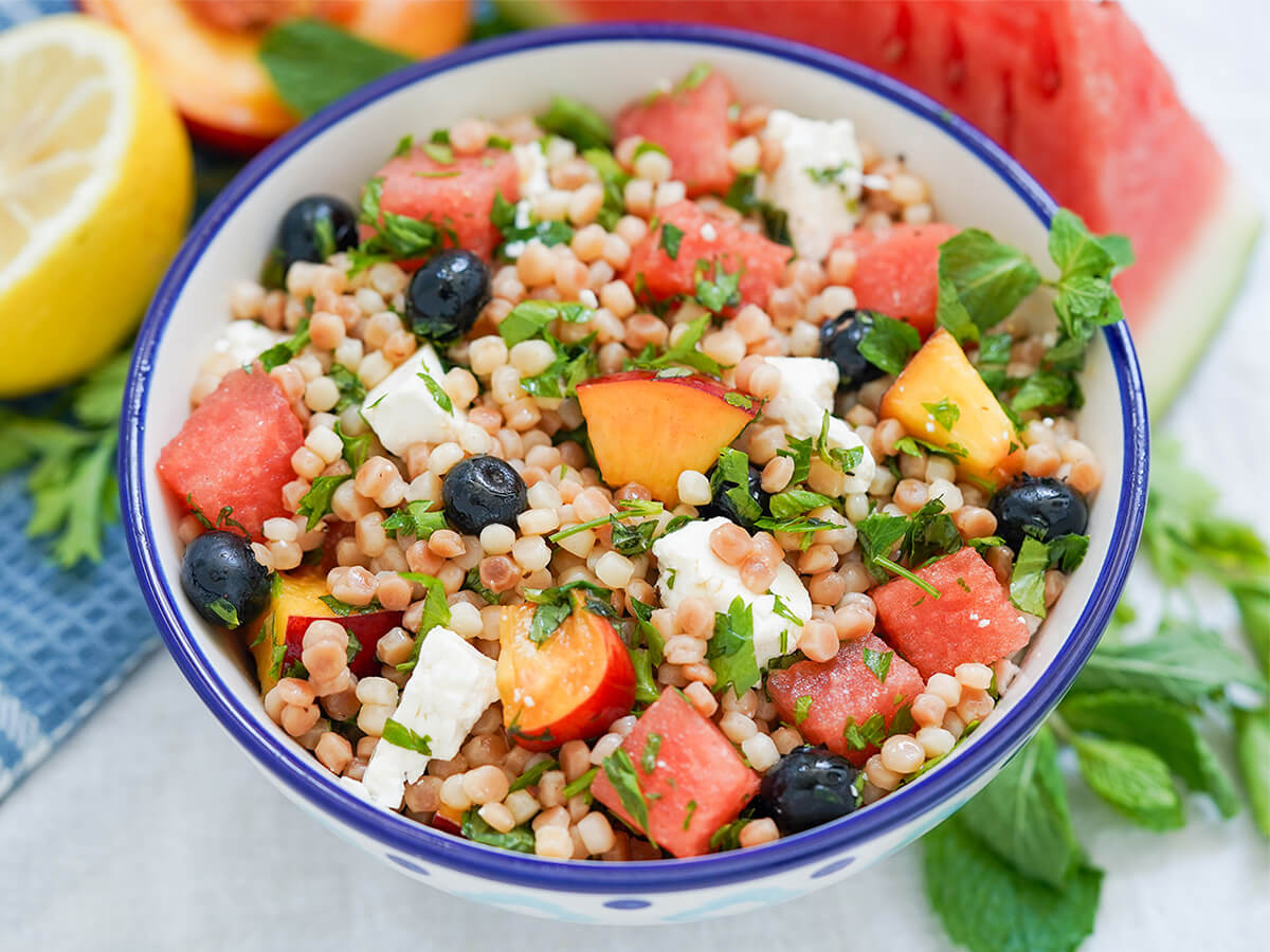 bowl of fruit couscous salad with herbs and feta bowl of fruit couscous salad with herbs around and watermelon, lemon and nectarine blurred behind.