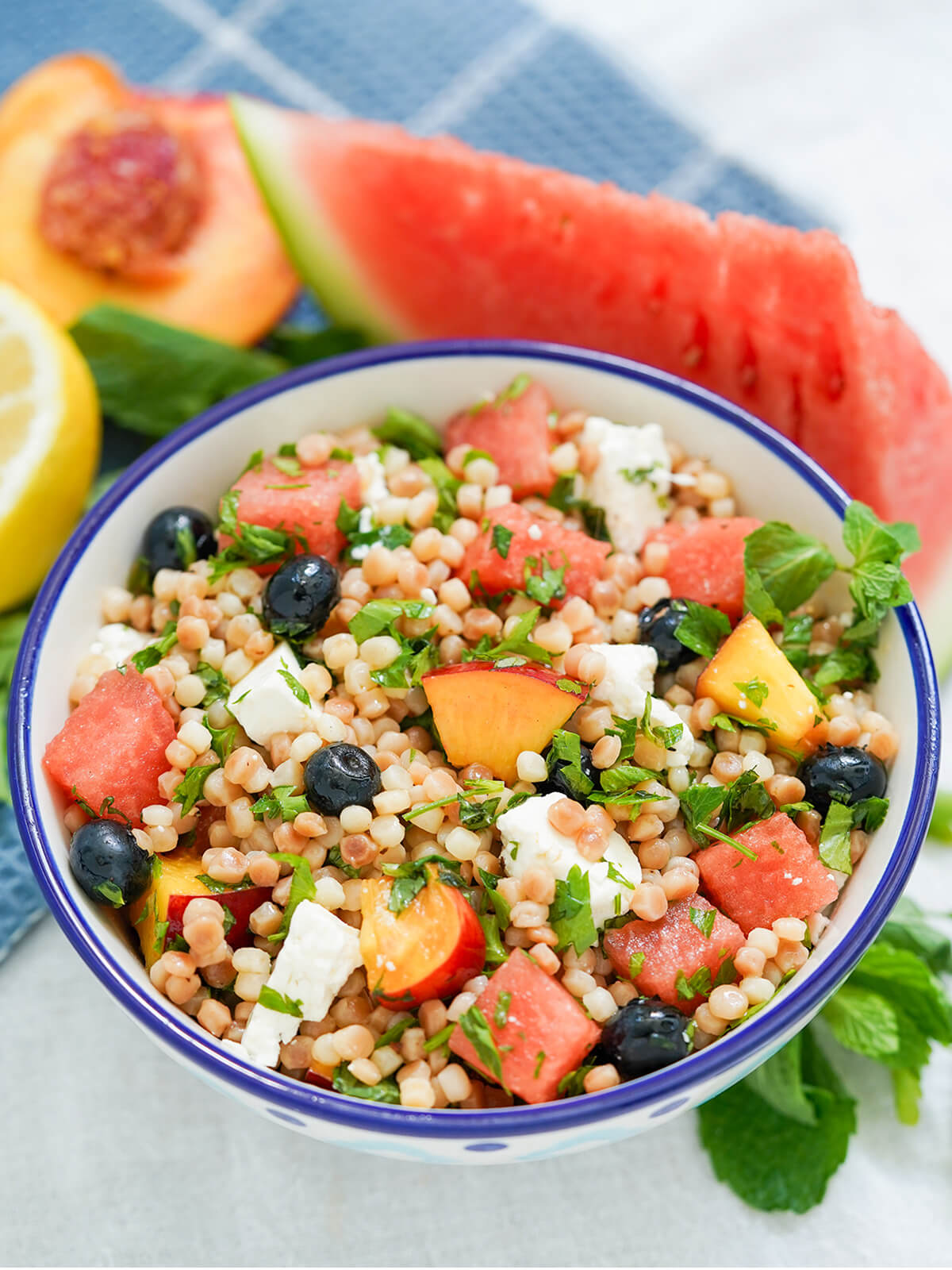 This fruit couscous salad with herbs and feta is light, bright and refreshing with juicy fresh fruit & aromatic herbs, but filling enough to be a great lunch or side. bowl of fruit couscous salad with wedge of watermelon and cut nectarine behind bowl.
