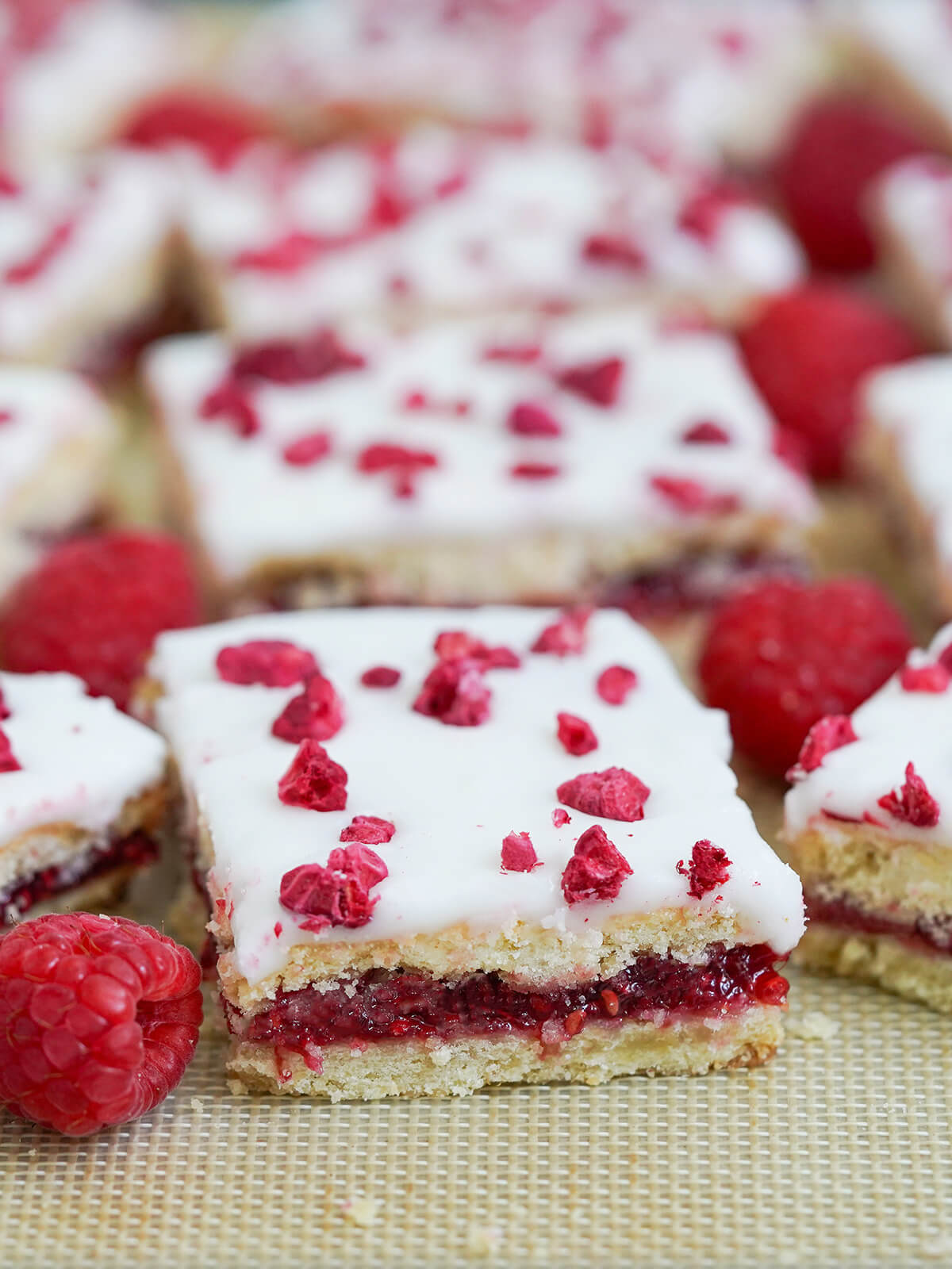 a slice of hindbaersnitter, Danish raspberry bars, with raspberries around and more slices blurred behind.