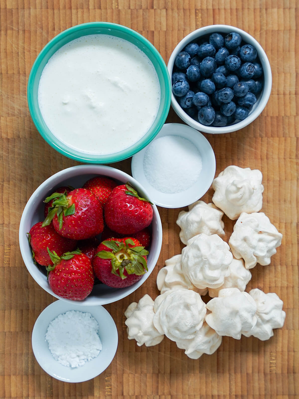 ingredients for Eton mess bowls of strawberries, blueberries, cream with meringues to side and small dishes with sugar.