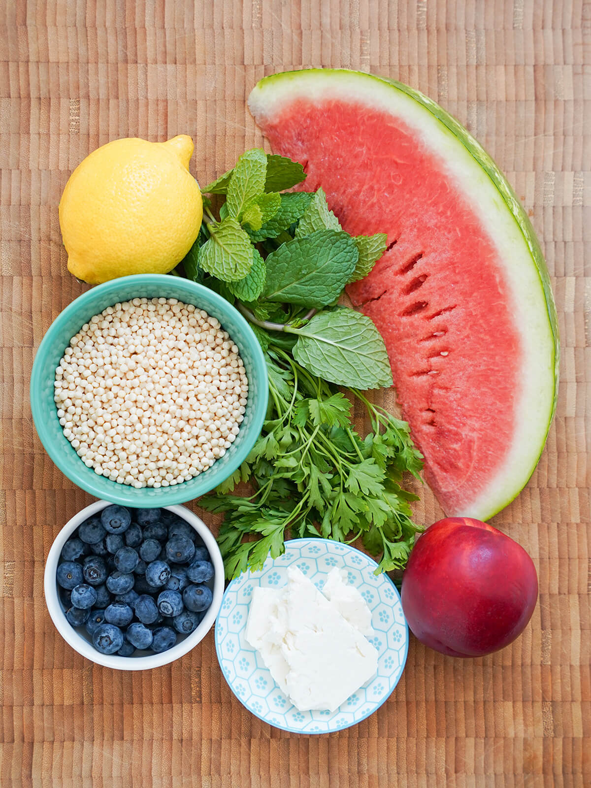 ingredients for fruit couscous salad bowl of uncooked pearl couscous, bowl of blueberries, feta, with wedge of watermelon, nectarine, lemon and herbs next to them.