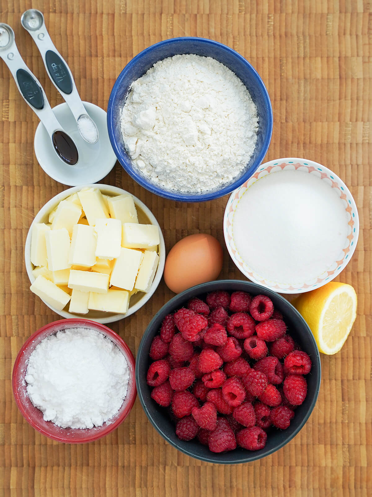 bowls of flour, sugar, raspberries, butter and powdered sugar with egg in middle and part lemon to side.