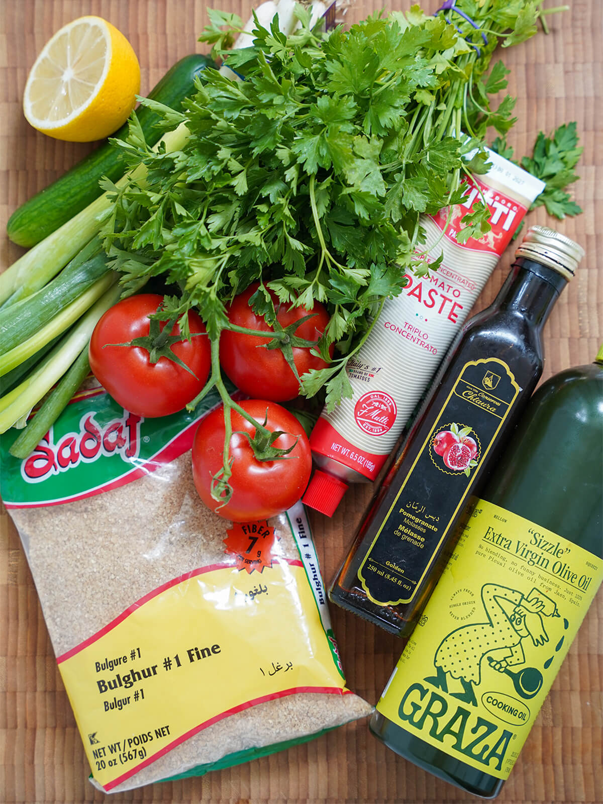 kisir ingredients bunch of parsley, tomatoes, lemon, cucumber, packet of fine bulgur, bottles of oil, pomegranate molasses and tomato paste on board.