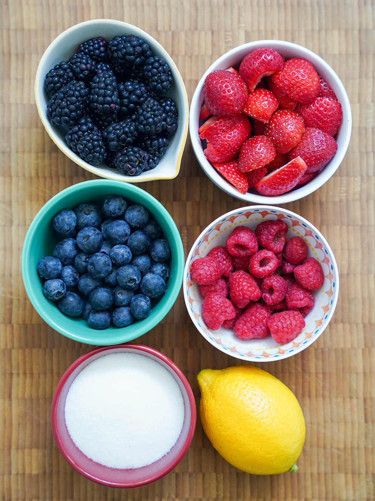bowls of blackberries, cut strawberries, raspberries and blueberries with bowl of sugar and lemon below.