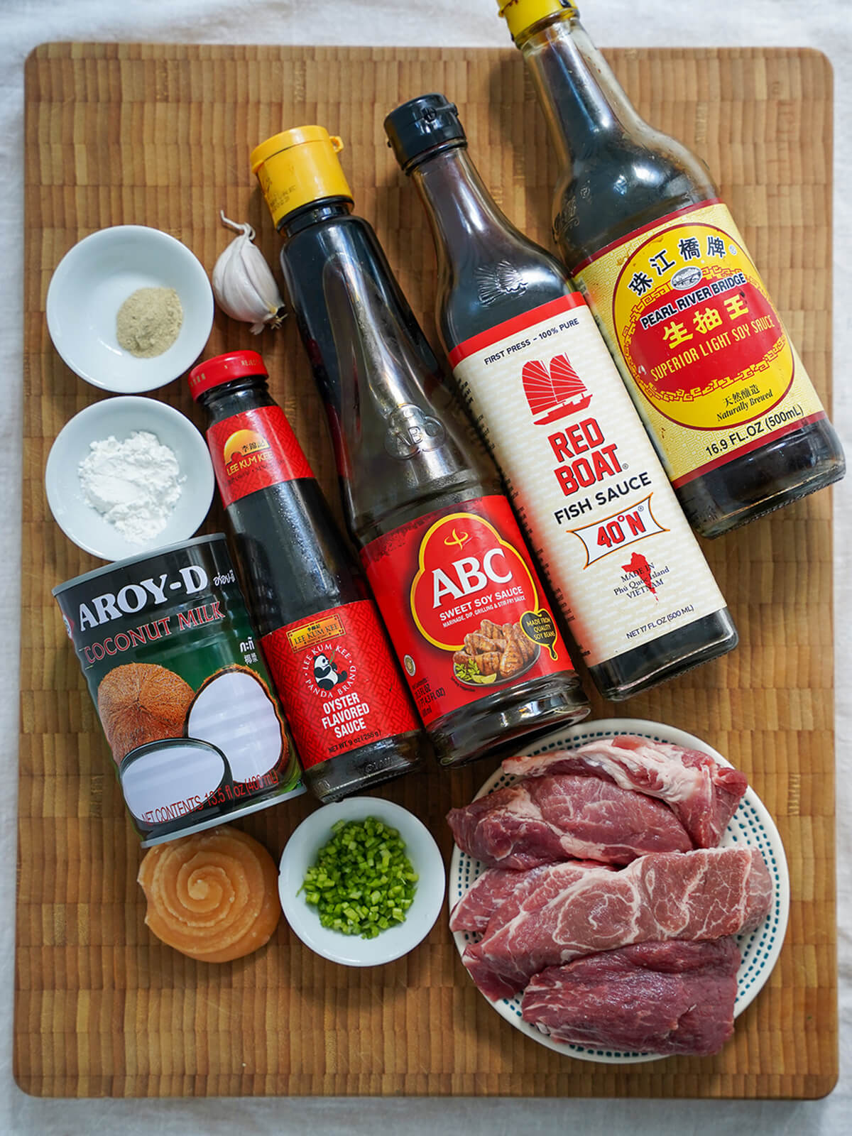 moo ping ingredients bottles of soy sauce, fish sauce, oyster sauce, coconut milk on board with pieces of pork, palm sugar, small dishes with chopped cilantro stem, white pepper and corn starch around.