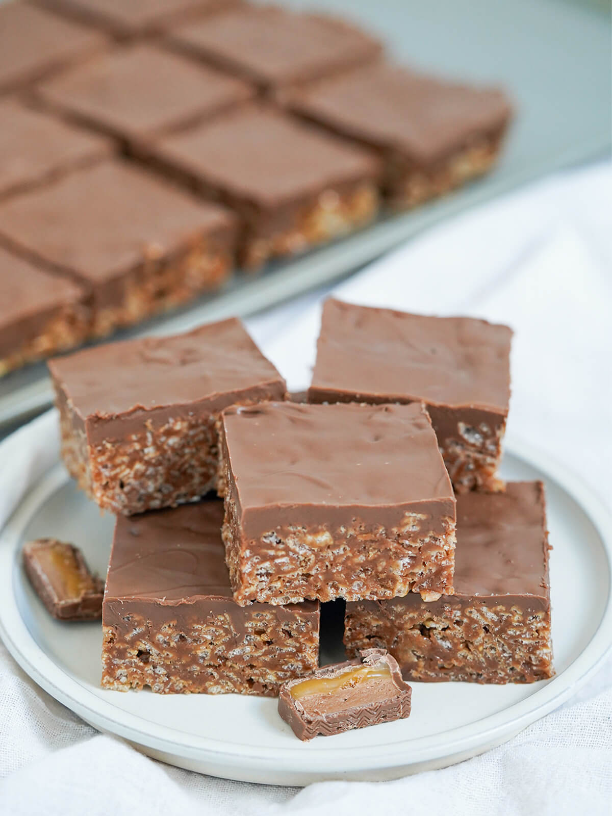 Mars bar slice is such an easy no bake treat that's quick to make with just a few ingredients. Always a favorite with all who try, perfect for parties & bake sales. plate with two layers of mars bar slice with more in rows blurred in background.