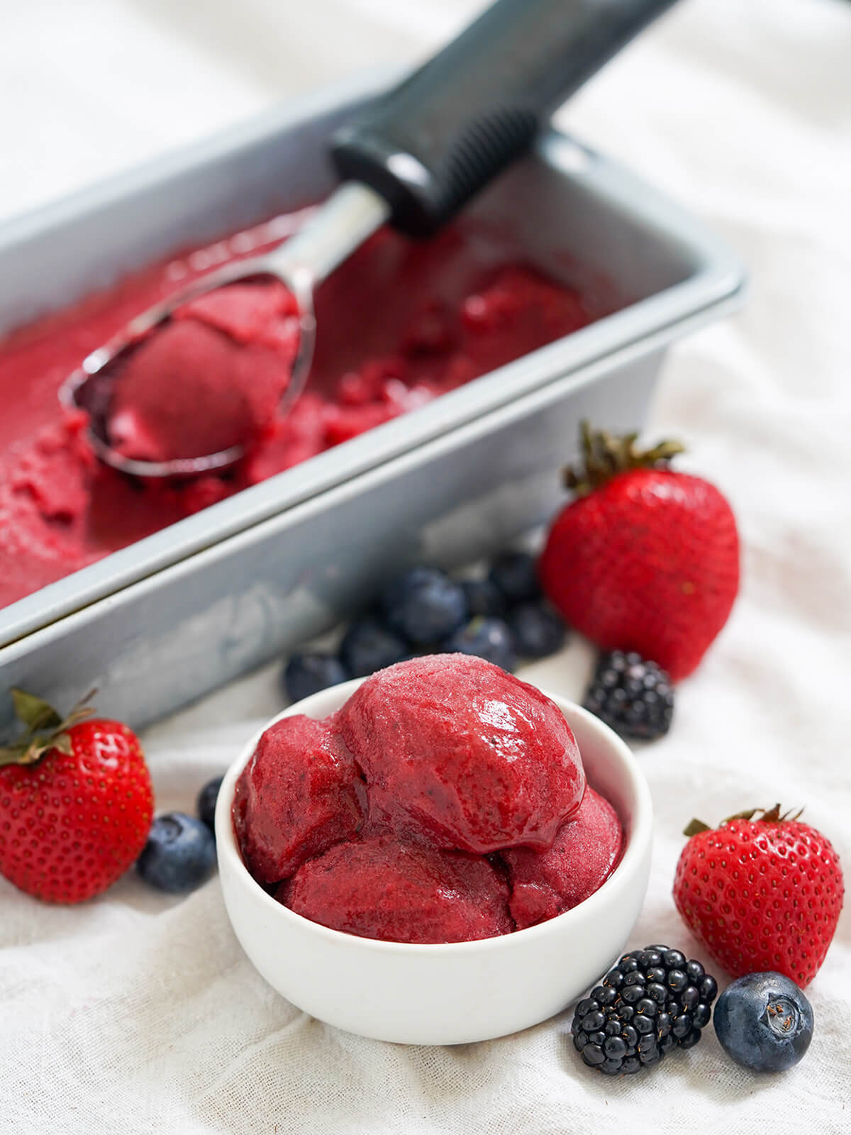 small bowl of mixed berry sorbet with berries around it and container of more and scoop resting on top of it behind.