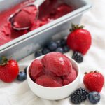 bowl of mixed berry sorbet with berries around it and scoop in container blurred in background.