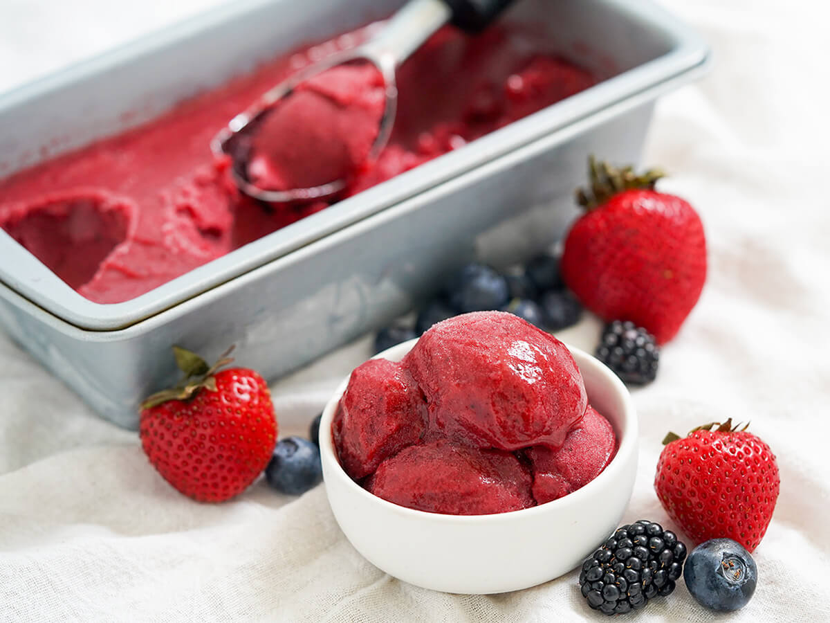 bowl of mixed berry sorbet with container with more behind, scoop resting on top, with berries around the bowl.